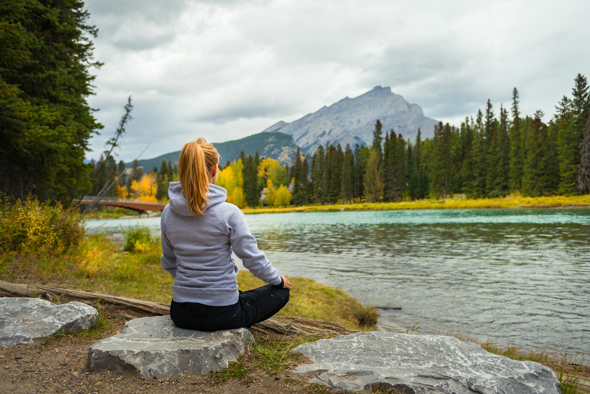 A woman sits crossed legged facing a lake / loch overlooking a pine forest, with a mountain behind it.